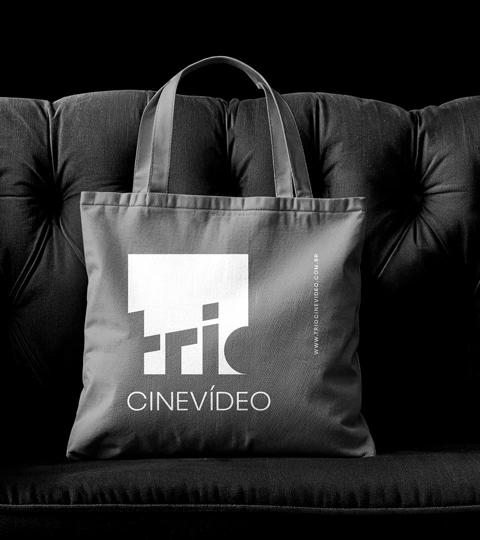 Ecobag cinza com a marca TRIO Cine Vídeo em branco, apoiada em um sofá preto de capitonê